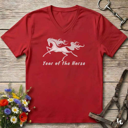 A red V-neck t-shirt laid flat on a rustic wooden surface, featuring a white graphic of two leaping horses and the text 'Year of the Horse'. The shirt is styled with equestrian tack, a small bouquet of colorful wildflowers, and metal horse bits.