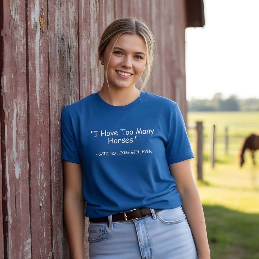 Woman wearing a blue t-shirt with text standing in front of a barn with a horse in the background. Never Too Many T-Shirt for Horse Lovers, Barn Wear, Riding Apparel Trail Riding Tee