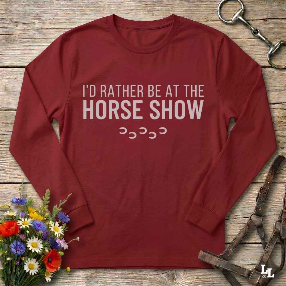 A red long-sleeve t-shirt with the text 'I'D RATHER BE AT THE HORSE SHOW' and a small stack of horseshoes printed on the front. The shirt is laid flat on a rustic wooden surface, styled with equestrian accessories (a leather bridle and metal tack) and a small bouquet of colorful wildflowers.