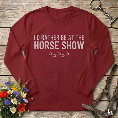 A red long-sleeve t-shirt with the text 'I'D RATHER BE AT THE HORSE SHOW' and a small stack of horseshoes printed on the front. The shirt is laid flat on a rustic wooden surface, styled with equestrian accessories (a leather bridle and metal tack) and a small bouquet of colorful wildflowers.