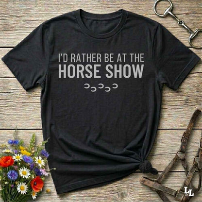 A black short-sleeve graphic T-shirt laid flat on a rustic wooden surface, with white text reading 'I'D RATHER BE AT THE HORSE SHOW' and small horse silhouettes underneath. The shirt is styled with flowers, a horse bit, and a leather bridle around it.