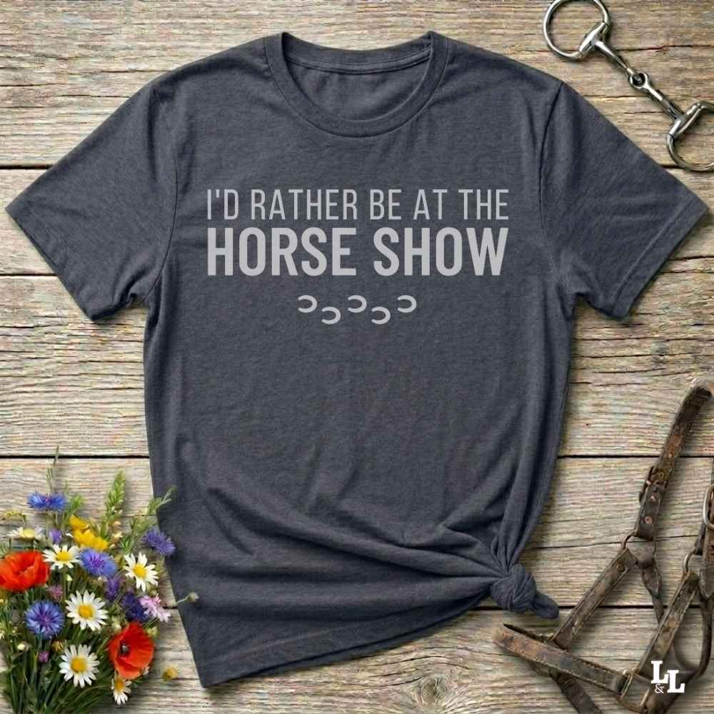A dark charcoal gray t-shirt with the text 'I'D RATHER BE AT THE HORSE SHOW' in white, laid flat on a rustic wooden background with decorative flowers and horse tack.