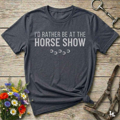 A dark charcoal gray t-shirt with the text 'I'D RATHER BE AT THE HORSE SHOW' in white, laid flat on a rustic wooden background with decorative flowers and horse tack.