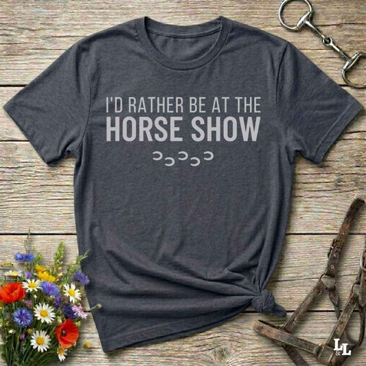A dark charcoal gray t-shirt with the text 'I'D RATHER BE AT THE HORSE SHOW' in white, laid flat on a rustic wooden background with decorative flowers and horse tack.