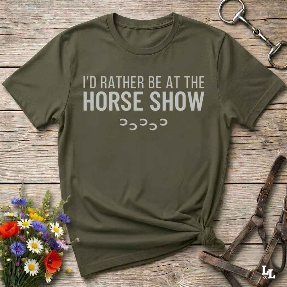 An olive green short-sleeve t-shirt with the text 'I'D RATHER BE AT THE HORSE SHOW' in white, laid flat on a rustic wooden surface with equestrian-themed props like a halter, horse bit, and wildflowers.