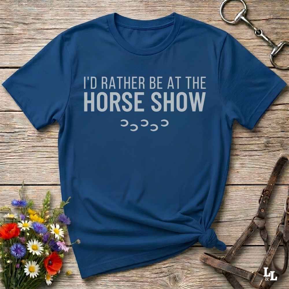 A navy blue graphic t-shirt with the text 'I'D RATHER BE AT THE HORSE SHOW' in white, styled in a flat-lay on a rustic wooden background with equestrian and floral props.