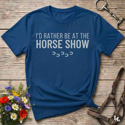 A navy blue graphic t-shirt with the text 'I'D RATHER BE AT THE HORSE SHOW' in white, styled in a flat-lay on a rustic wooden background with equestrian and floral props.