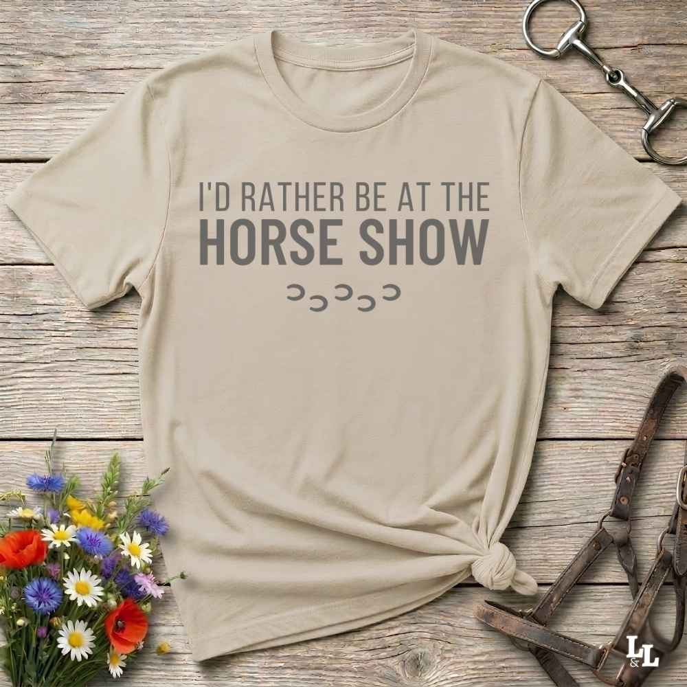 A beige graphic t-shirt with the text 'I'D RATHER BE AT THE HORSE SHOW' and hoof prints, styled in a flat-lay on a rustic wooden background with equestrian accessories and flowers.