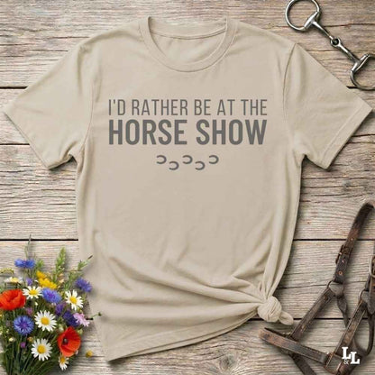 A beige graphic t-shirt with the text 'I'D RATHER BE AT THE HORSE SHOW' and hoof prints, styled in a flat-lay on a rustic wooden background with equestrian accessories and flowers.