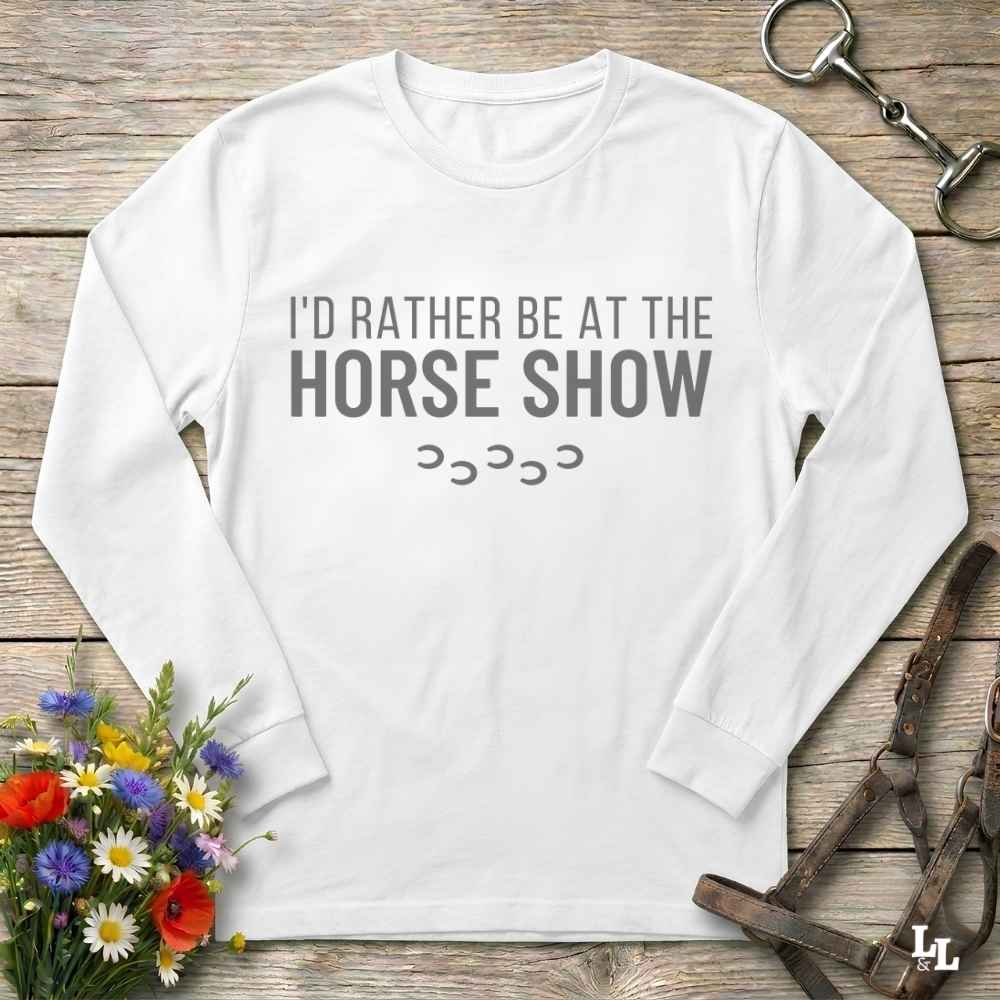 A white long-sleeve t-shirt with the text 'I'D RATHER BE AT THE HORSE SHOW' and a small row of horseshoes printed on the front, displayed flat on a rustic wooden background with equestrian accessories and flowers.