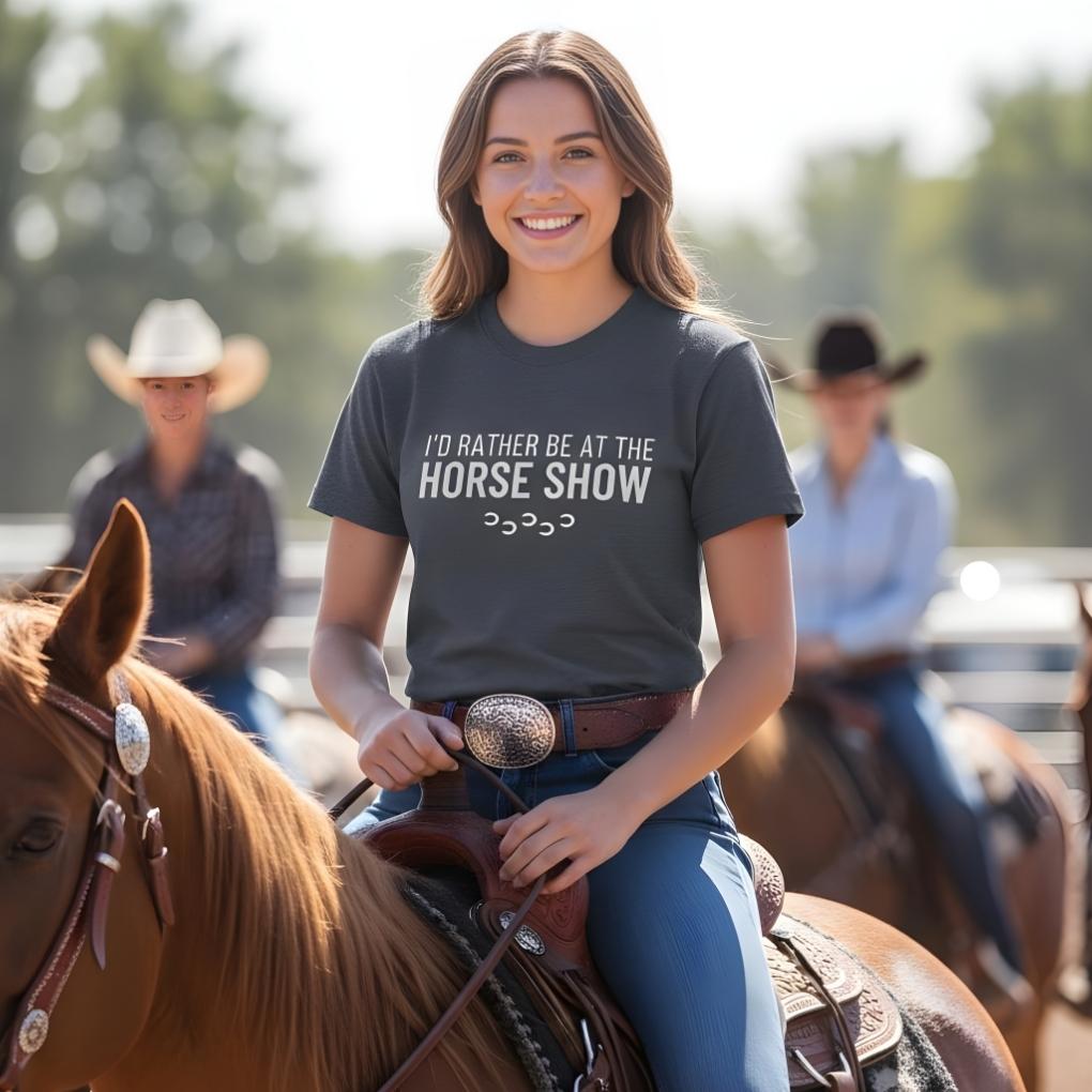 Woman riding a horse wearing a t-shirt with a horse show-related message, with blurred background of other riders. I'd Rather be at the Horse Show T-Shirt for Horse Riders, Trail Riding Tee