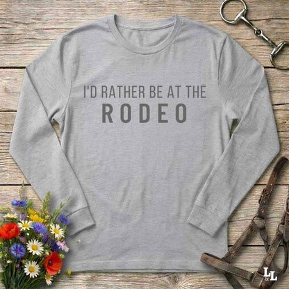 Grey long-sleeve shirt with 'I'd Rather Be at the Rodeo' text on a wooden surface with floral and equestrian-themed accessories.