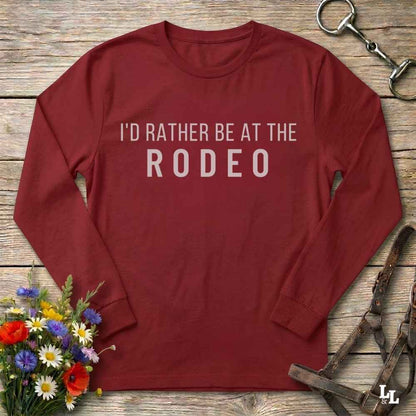A long-sleeve crewneck shirt in dark red with the text 'I'D RATHER BE AT THE RODEO' printed in white across the chest. The shirt is laid flat on a rustic wooden surface, surrounded by equestrian-themed props including a bit, horse bits, and a small bouquet of wildflowers.