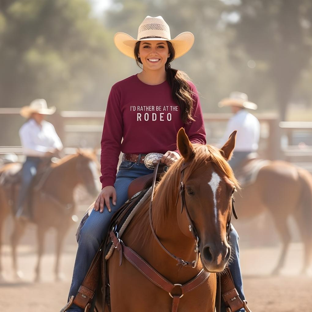 Woman riding a horse wearing a shirt that says 'I'd Rather Be at the Rodeo' with a blurred background of other riders. I'd Rather be a the Rodeo Long Sleeve Tee for Horse Riders, Equestrian Apparel. Perfect gift for horse people.