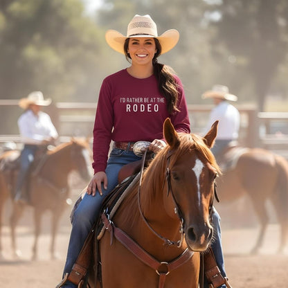 Woman riding a horse wearing a shirt that says 'I'd Rather Be at the Rodeo' with a blurred background of other riders. I'd Rather be a the Rodeo Long Sleeve Tee for Horse Riders, Equestrian Apparel. Perfect gift for horse people.