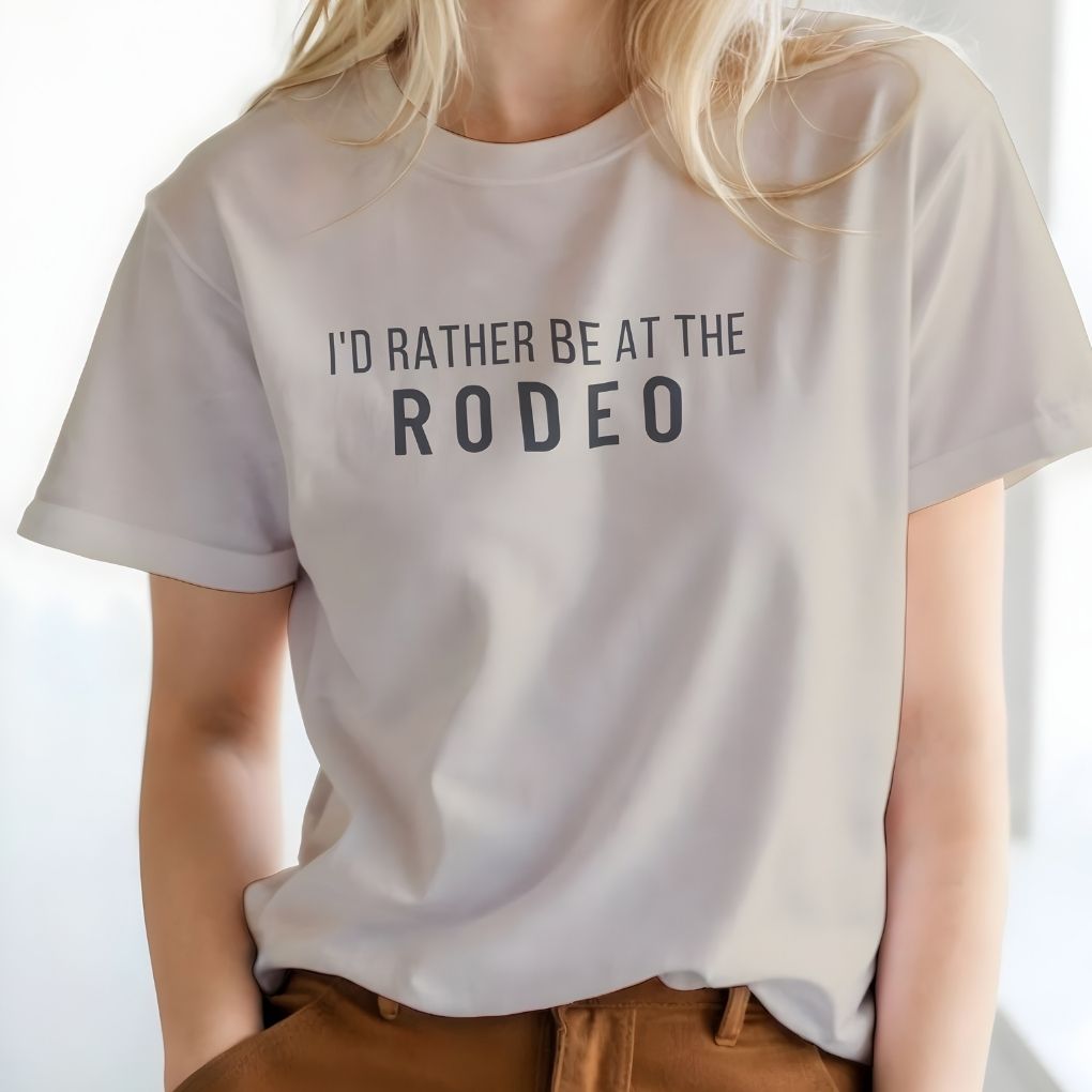 Person wearing a white t-shirt with 'I'd Rather Be at the Rodeo' text on a plain background. I'd Rather be at the Rodeo T-Shirt for Horse Lovers, Trail Riding Tee Barn Wear perfect for a gift