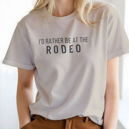Person wearing a white t-shirt with 'I'd Rather Be at the Rodeo' text on a plain background. I'd Rather be at the Rodeo T-Shirt for Horse Lovers, Trail Riding Tee Barn Wear perfect for a gift