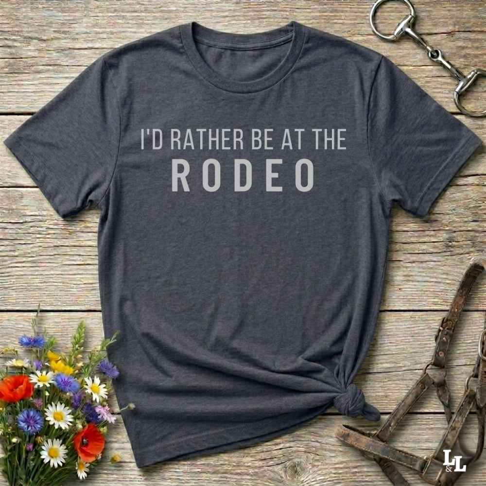 A dark charcoal gray graphic t-shirt with the text 'I'D RATHER BE AT THE RODEO' in white, styled in a flat-lay on a rustic wooden background with equestrian accessories and flowers.