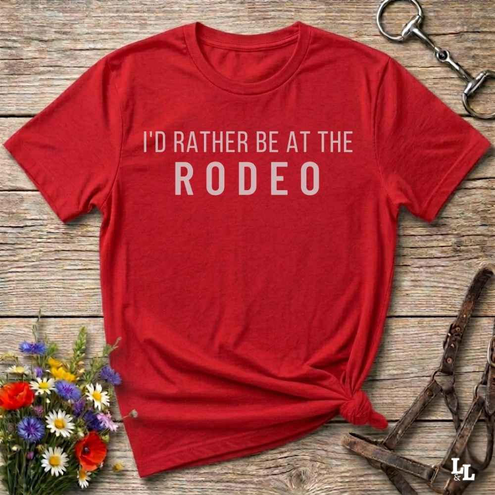 A red short-sleeve t-shirt with the text 'I'D RATHER BE AT THE RODEO' printed in white across the chest, styled in a flat lay on a rustic wooden background with equestrian and floral props.