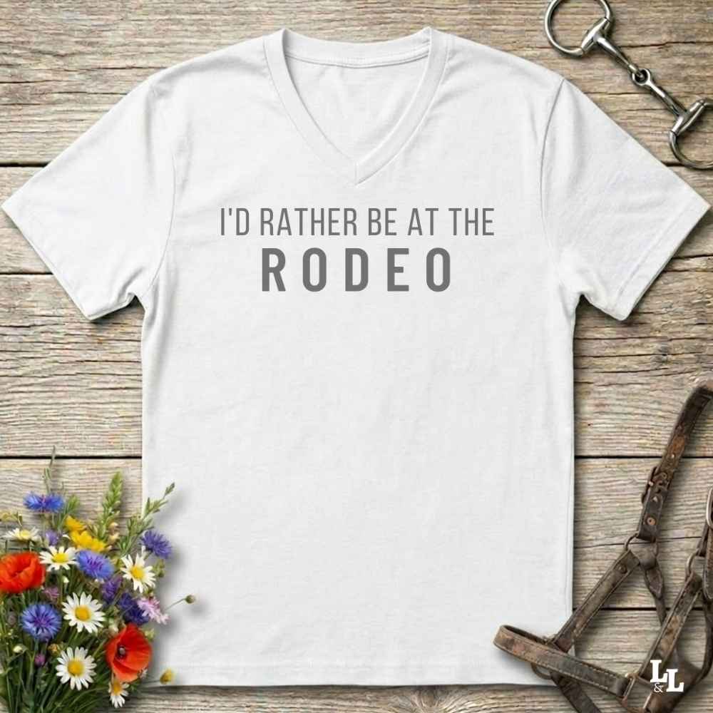 A white V-neck t-shirt with the text 'I'D RATHER BE AT THE RODEO' printed in white across the chest, styled in a flat lay on a rustic wooden background with equestrian accessories and flowers.