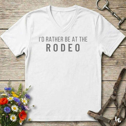 A white V-neck t-shirt with the text 'I'D RATHER BE AT THE RODEO' printed in white across the chest, styled in a flat lay on a rustic wooden background with equestrian accessories and flowers.