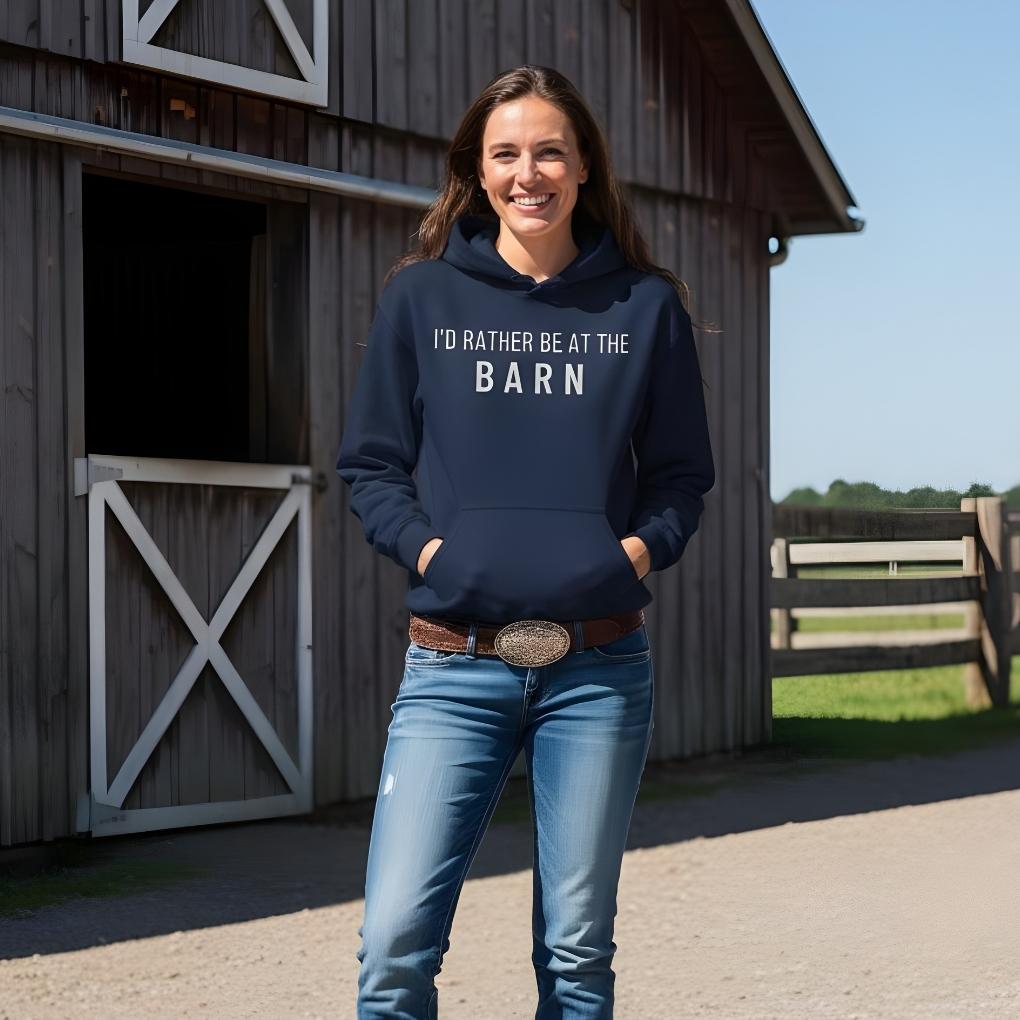 Woman wearing a navy hoodie with 'I'd Rather Be at the Barn' text, standing in front of a wooden barn. I'd Rather be at the Barn Hoodie for Horse Riders, Barn Wear Hoodie, Equestrian Apparel. Great gift for horse lover.