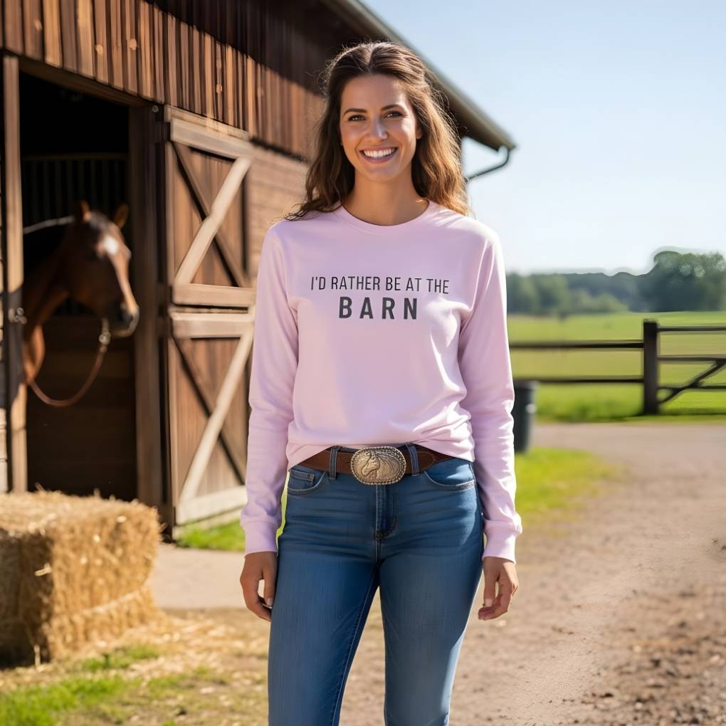 Woman wearing a pink sweatshirt with 'I'd Rather Be at the Barn' text, standing in front of a barn with a horse. I'd Rather be at the Barn Long Sleeve Tee for Horse Lovers, Barn Wear, Equestrian Tee. Great gift for horse riders.