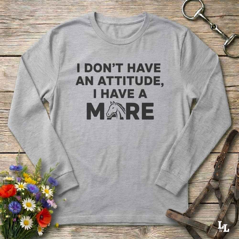 A light gray long-sleeve t-shirt with a black graphic text print reading 'I DON'T HAVE AN ATTITUDE, I HAVE A MORE' with a horse head silhouette, displayed on a rustic wooden background with equestrian accessories and flowers.