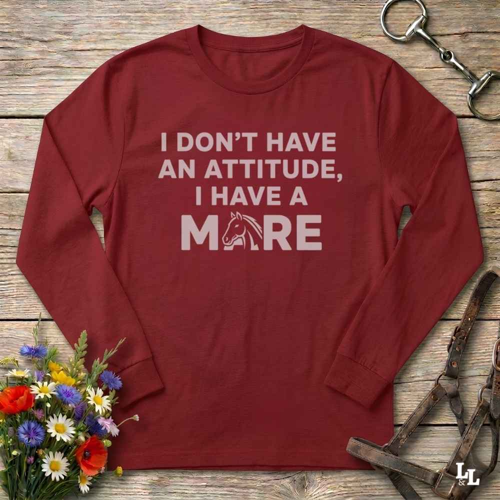 A long-sleeve crewneck t-shirt in maroon red with a white graphic text that reads 'I DON'T HAVE AN ATTITUDE, I HAVE A MARE' with a horse head silhouette inside the letter 'A'. The shirt is displayed flat on a rustic wooden background with equestrian accessories and flowers.