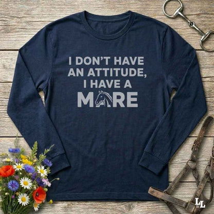 A navy blue long-sleeve t-shirt with a white graphic text print that reads 'I DON'T HAVE AN ATTITUDE, I HAVE A MARE' with a horse silhouette inside the letter 'M'. The shirt is displayed in a flat-lay arrangement on a rustic wooden background, surrounded by equestrian-themed props including a horse bit, leather bridle, and a small bouquet of wildflowers.
