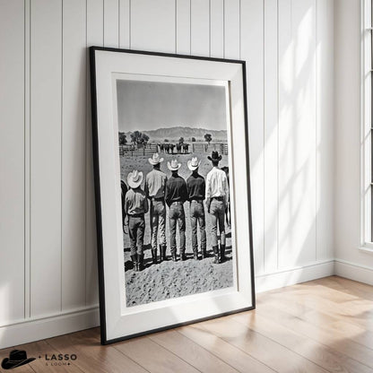 Framed black and white photograph of cowboys in a line, leaning against a fence, in a room with wooden flooring and white walls.
