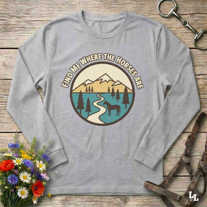 A light gray long-sleeve t-shirt with a circular graphic on the chest that reads 'FIND ME WHERE THE HORSES ARE' above a mountain, lake, river, and horse illustration. The shirt is laid flat on a rustic wooden surface with decorative props including flowers, a horse bit, and a leather bridle.