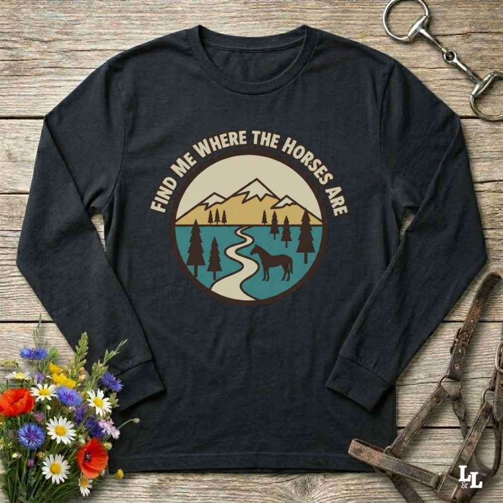 A black long-sleeve t-shirt with a circular graphic print on the chest, laid flat on a rustic wooden surface with decorative flowers and horse tack props.
