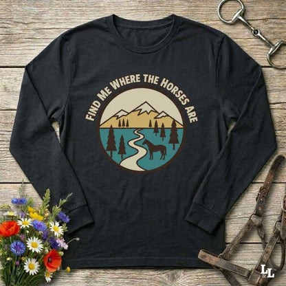 A black long-sleeve t-shirt with a circular graphic print on the chest, laid flat on a rustic wooden surface with decorative flowers and horse tack props.