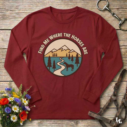 A long-sleeve crewneck t-shirt in maroon red, laid flat on a rustic wooden background. The shirt features a circular graphic in the center with the text 'FIND ME WHERE THE HORSES ARE' arched over a retro-style mountain, lake, and horse illustration.
