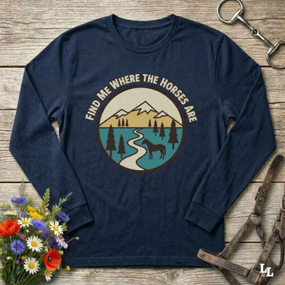 A navy blue long-sleeve t-shirt with a circular graphic on the chest that reads 'FIND ME WHERE THE HORSES ARE' above a mountain, lake, and horse illustration. The shirt is displayed in a flat-lay arrangement on a rustic wooden background, accompanied by flowers, a horse bit, and a leather bridle.