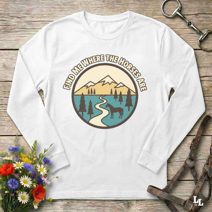 A white long-sleeve crewneck t-shirt with a circular graphic print on the chest. The graphic features a mountain landscape with a winding river, trees, and a horse silhouette, with the text 'FIND ME WHERE THE HORSES ARE' arched above.
