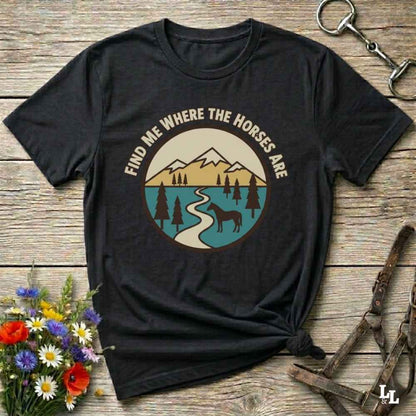 A black graphic t-shirt laid flat on a rustic wooden surface, styled with a small bouquet of flowers, a horse bit, and a leather halter. The t-shirt features a circular graphic with the text 'FIND ME WHERE THE HORSES ARE' surrounding a mountain, river, and horse silhouette design.