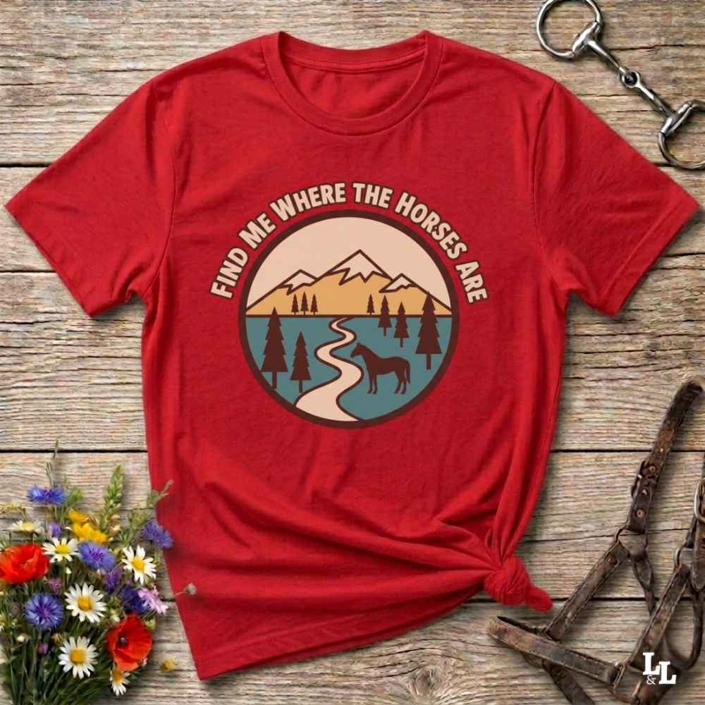 A red short-sleeve graphic T-shirt laid flat on a rustic wood surface, with a circular mountain-and-lake print and the text 'FIND ME WHERE THE HORSES ARE' across the chest. Decorative flowers and horse tack are placed around it as styling props.