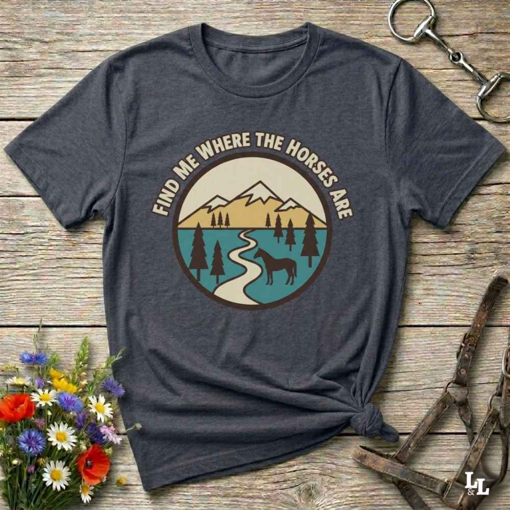 A dark heather gray graphic t-shirt laid flat on a rustic wooden surface, styled with a horse halter, flowers, and a small horse head icon. The t-shirt features a circular graphic of mountains, a lake, pine trees, and a horse silhouette with the text 'FIND ME WHERE THE HORSES ARE' arched above.