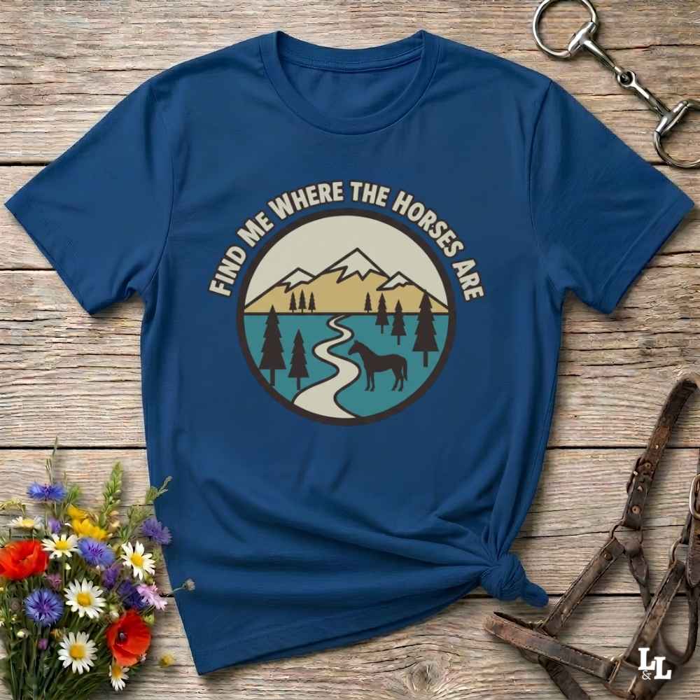 A navy blue graphic t-shirt laid flat on a rustic wooden surface, styled with a small bouquet of flowers, a metal horse bit, and a brown leather halter. The t-shirt features a circular graphic with the text 'FIND ME WHERE THE HORSES ARE' surrounding an illustration of mountains, a river, pine trees, and a horse silhouette.