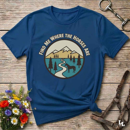 A navy blue graphic t-shirt laid flat on a rustic wooden surface, styled with a small bouquet of flowers, a metal horse bit, and a brown leather halter. The t-shirt features a circular graphic with the text 'FIND ME WHERE THE HORSES ARE' surrounding an illustration of mountains, a river, pine trees, and a horse silhouette.