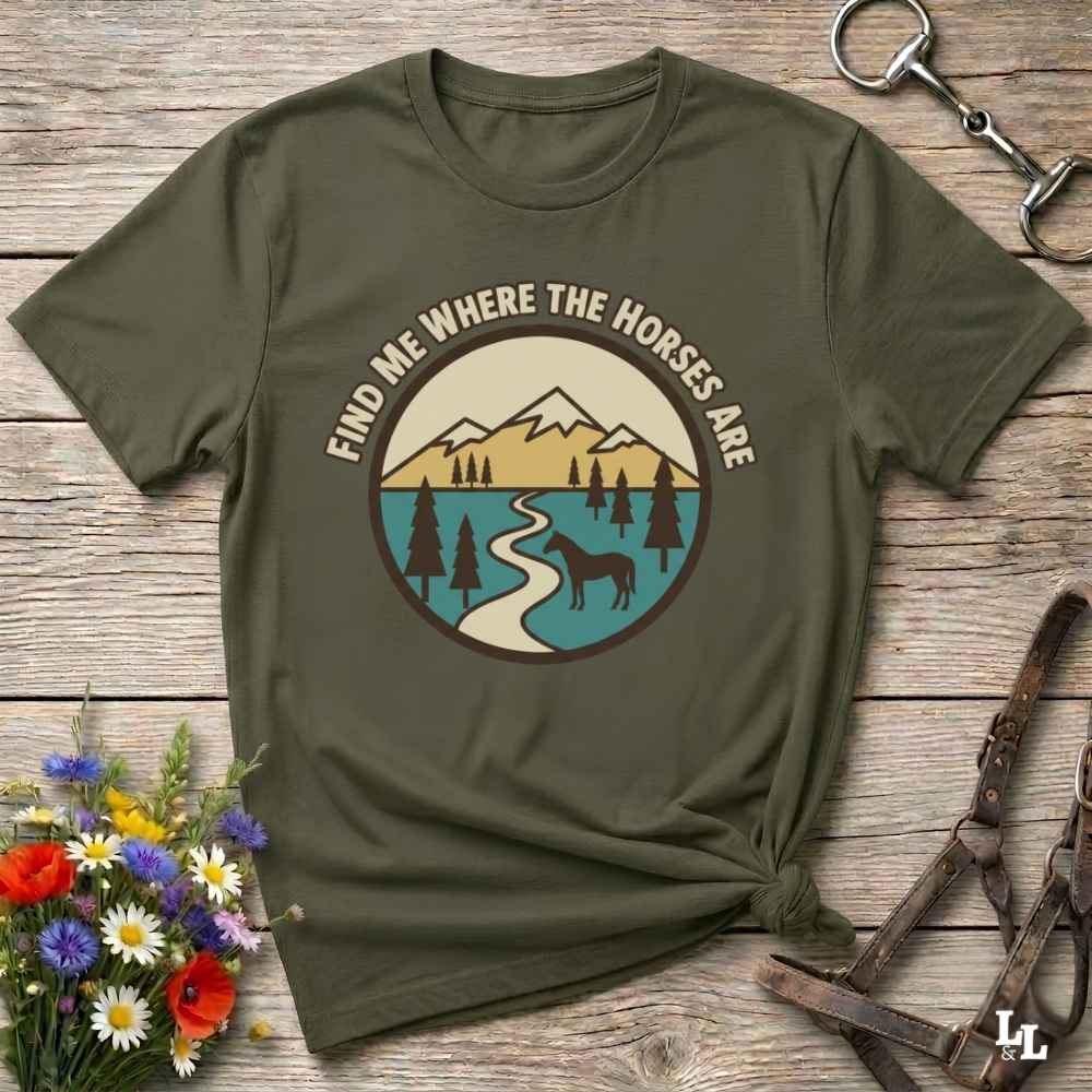 An olive green graphic t-shirt laid flat on a rustic wooden surface, styled with equestrian accessories and a small bouquet of wildflowers. The t-shirt features a circular graphic with the text 'FIND ME WHERE THE HORSES ARE' surrounding an illustration of mountains, a river, pine trees, and a horse silhouette.