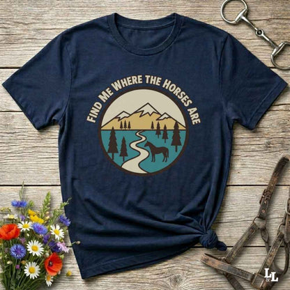 A navy blue graphic t-shirt laid flat on a rustic wooden surface, styled with equestrian accessories and a small bouquet of wildflowers. The t-shirt features a circular retro-style graphic of mountains, a river, and a horse, with the text 'FIND ME WHERE THE HORSES ARE' arched above the illustration.