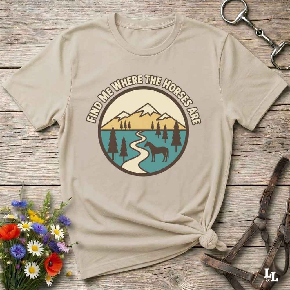 A beige graphic t-shirt with a circular mountain, lake, and horse design, styled on a rustic wooden background with flowers and leather tack.