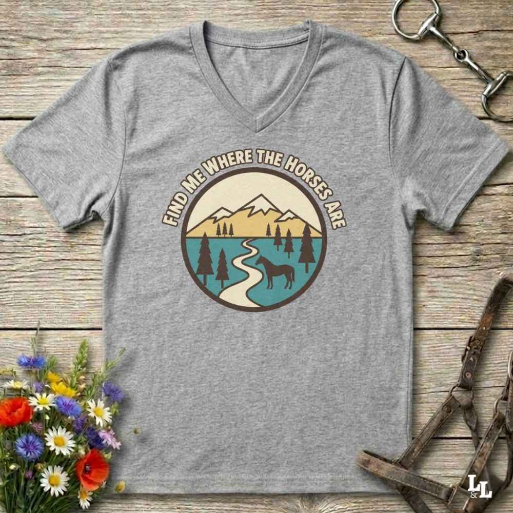 A heather gray V-neck t-shirt laid flat on a rustic wooden surface, featuring a circular graphic with mountains, trees, a winding river, and a horse silhouette, with the text 'FIND ME WHERE THE HORSES ARE' around the design. Decorative flowers and horse tack are placed around the shirt.