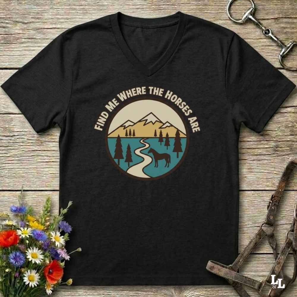 A black V-neck t-shirt with a circular graphic of a mountain landscape, river, pine trees, and a horse, with the text 'FIND ME WHERE THE HORSES ARE' arched above it. The shirt is displayed flat on a rustic wooden background with equestrian accessories and flowers.