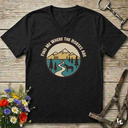 A black V-neck t-shirt with a circular graphic of a mountain landscape, river, pine trees, and a horse, with the text 'FIND ME WHERE THE HORSES ARE' arched above it. The shirt is displayed flat on a rustic wooden background with equestrian accessories and flowers.