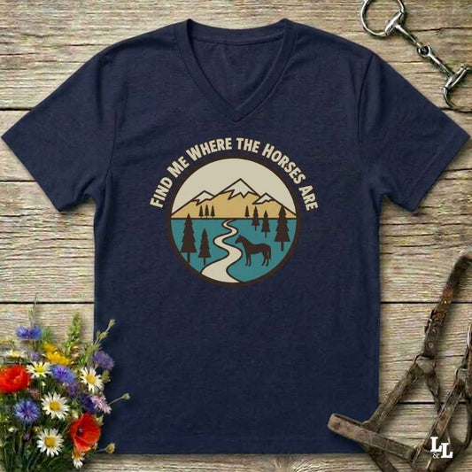 A navy blue V-neck t-shirt laid flat on a rustic wooden surface, featuring a circular graphic of a mountain landscape with a winding river and a horse silhouette, with the text 'FIND ME WHERE THE HORSES ARE' arched above. Decorative flowers and horse tack are placed around the shirt.