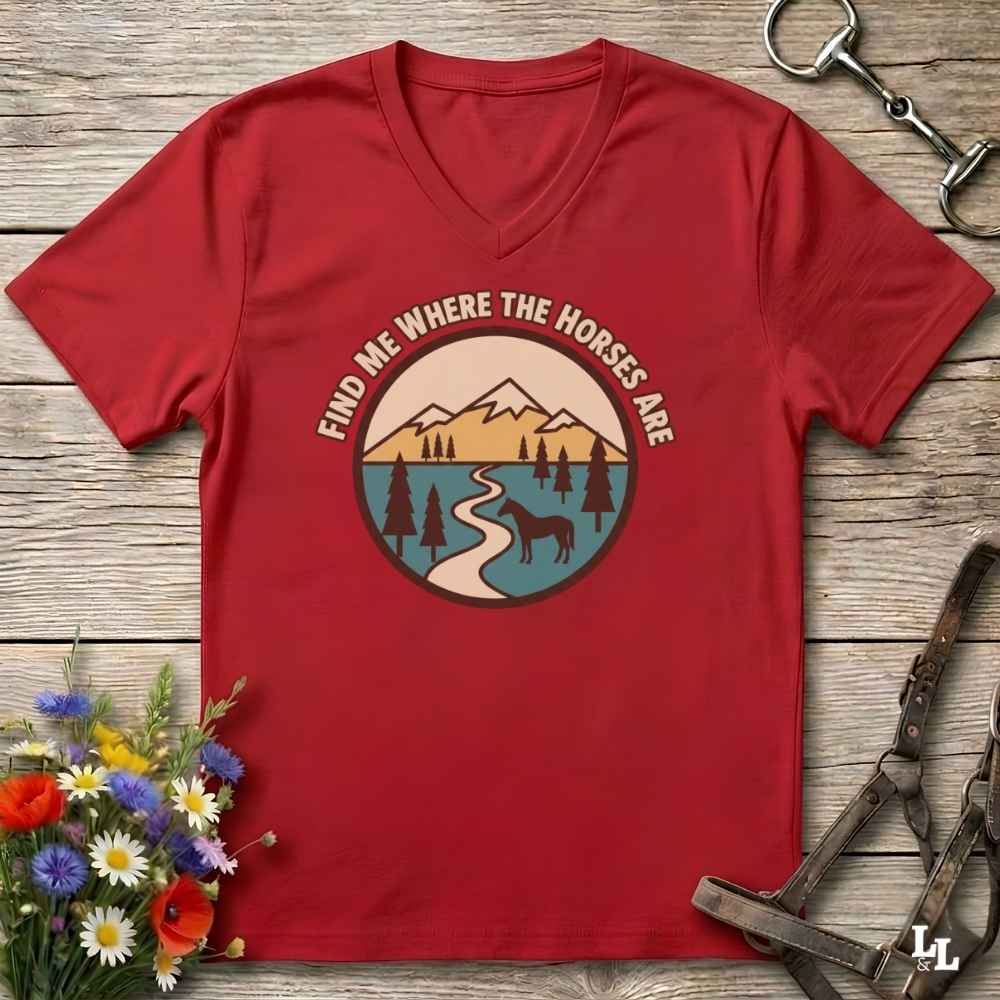 A red V-neck t-shirt with a circular graphic print featuring mountains, a river, pine trees, and a deer, with the text 'FIND ME WHERE THE HORSES ARE' arched above it. The shirt is displayed in a flat-lay arrangement on a rustic wooden background, surrounded by equestrian tack and a small bouquet of wildflowers.